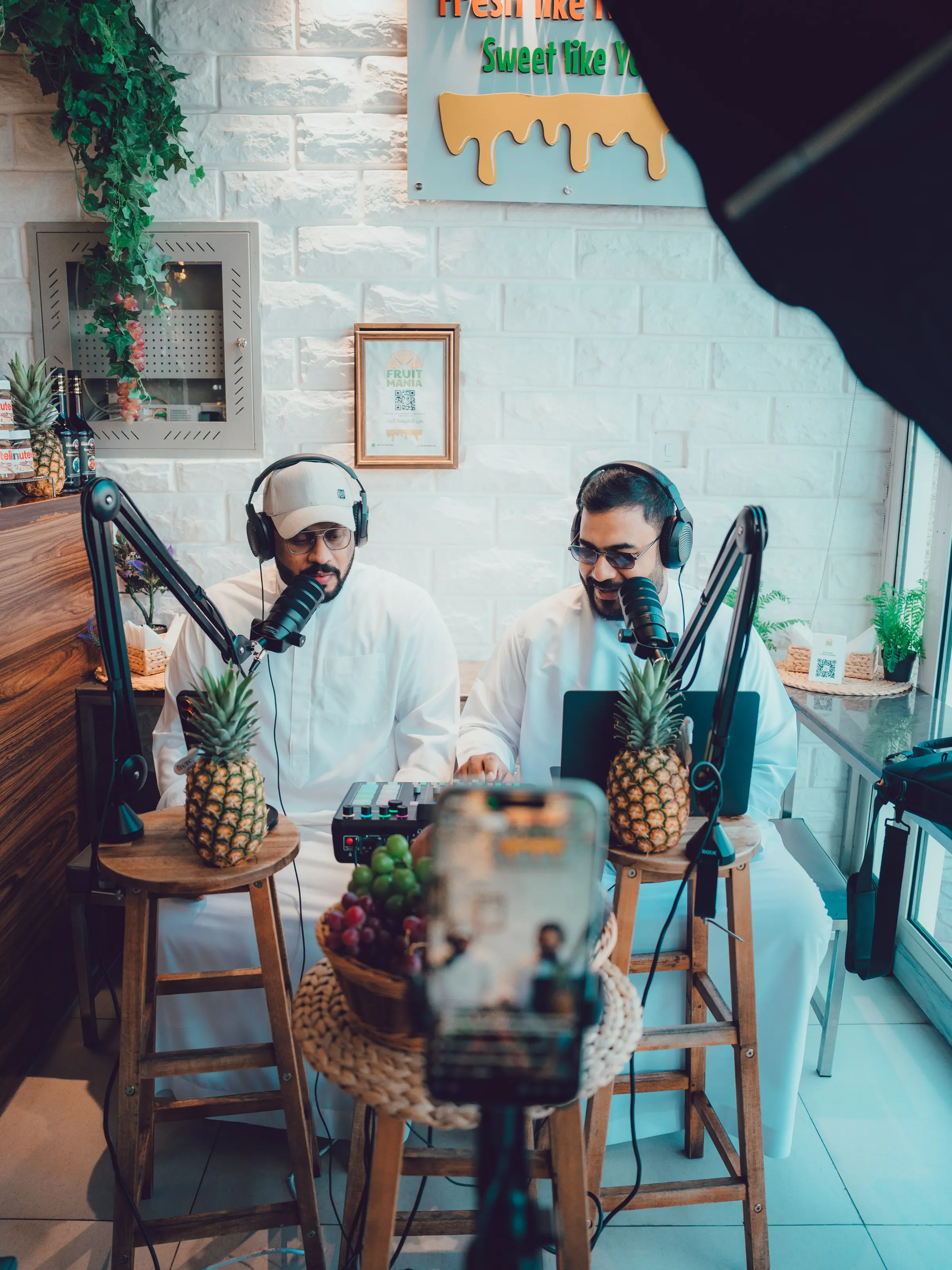 Two presenters in white kanduras seated at microphones on a set dressed with fruit and plants