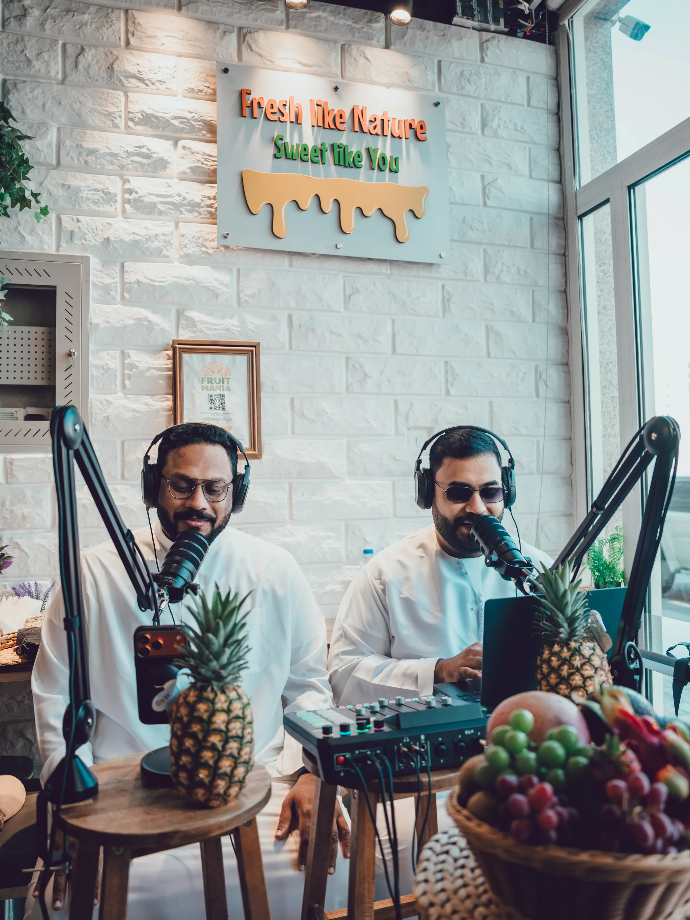 A wide view of a podcast recording in progress, two presenters at microphones with fruit and plants dressing the set