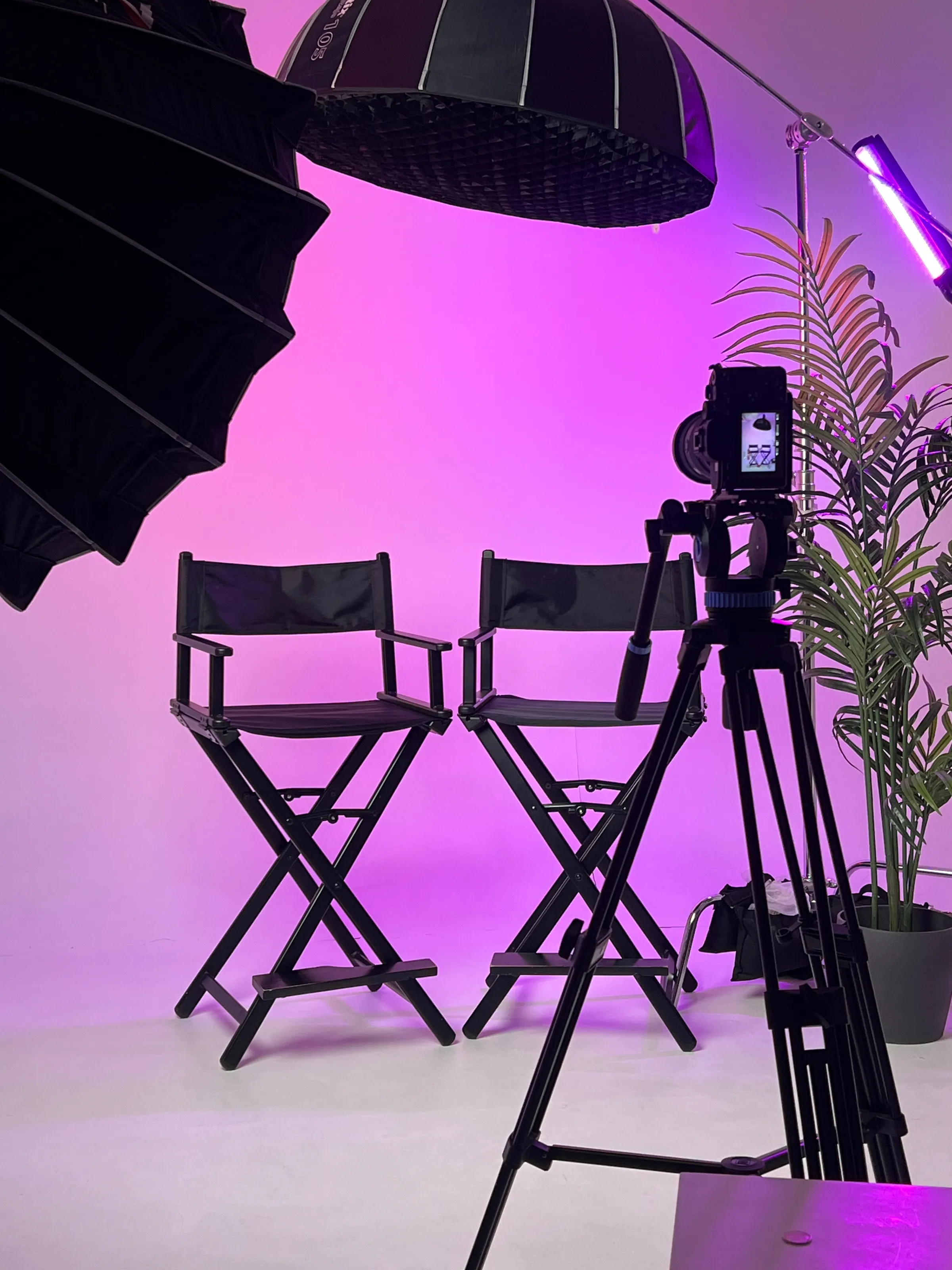 Two director's chairs facing a cinema camera on a tripod under magenta studio lighting